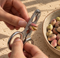 Hands using stainless steel pistachio opener pliers to crack a pistachio beside a bowl of pistachios